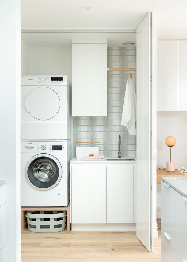 Bright, compact laundry room viewed through an open doorway. A stacked white washer and dryer sit beside white built-in cabinetry and a sink vanity. The rear wall features narrow vertical grey patterned tiles with timber rails, while pale timber flooring and white cabinetry create a clean, modern look. Part of an adjoining kitchen or living area is visible at right.