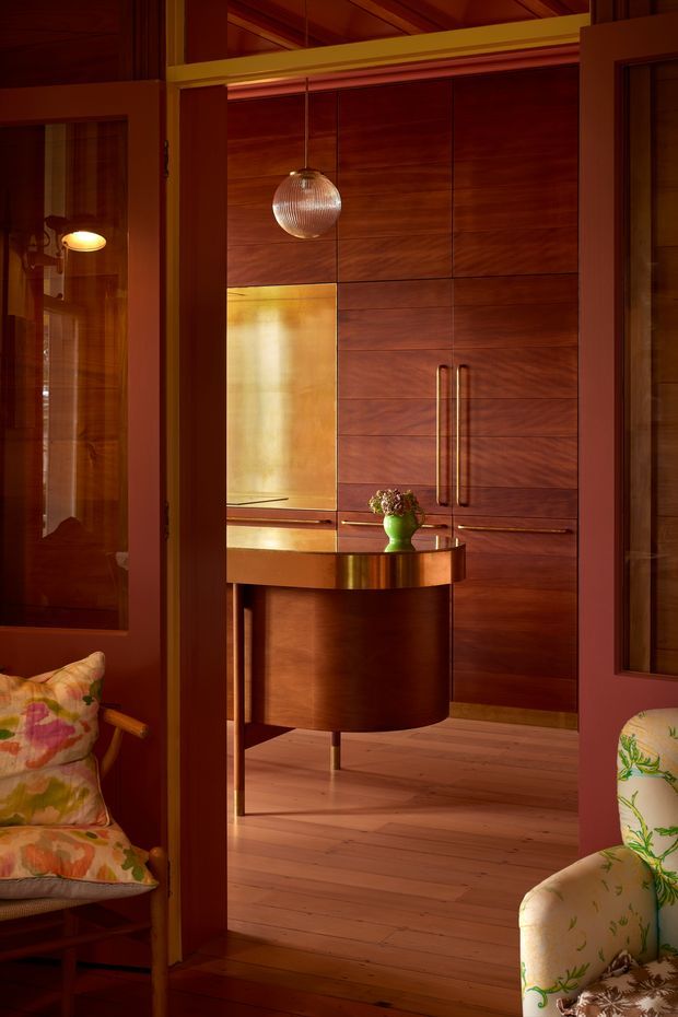 A warmly lit, richly detailed timber interior viewed through a doorway, featuring a wall of dark wood built-in cabinetry with long vertical handles, a curved dark wood console or bar cabinet, a brass-toned inset panel, a ribbed glass pendant light, and wide timber flooring. Upholstered floral seating is visible in the foreground on either side, with timber-framed glazed openings and exposed timber ceiling elements contributing to the refined, mid-century-inspired atmosphere.