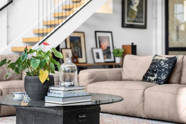 A bright contemporary living room with a large beige sectional sofa, black rectangular coffee table, potted plant, stacked books, framed artwork, and a staircase with timber treads and slender dark railing. The room has white walls, soft natural light, and a relaxed, curated feel.