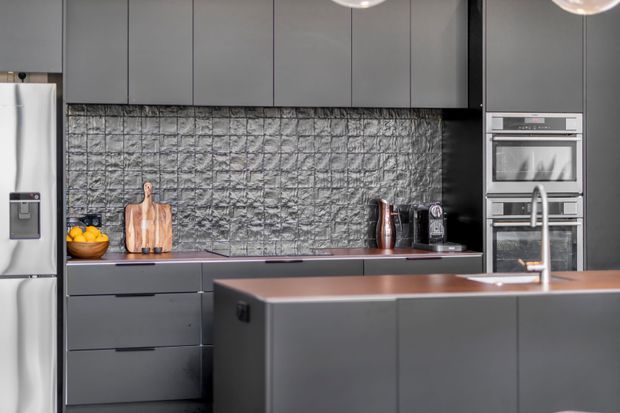 A contemporary dark-toned kitchen with flat-panel charcoal cabinetry, a large central island, integrated stainless-steel appliances, and a dramatic textured grey stone-look splashback. Copper-toned benchtops and pendant lights add warmth, while timber chopping boards and small countertop items provide subtle detail.