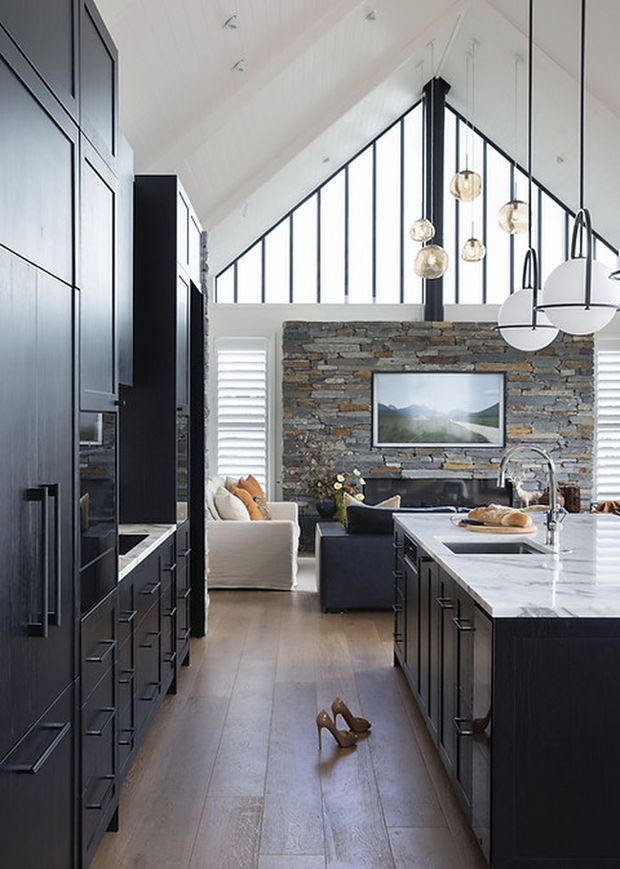A bright open-plan kitchen and living area with a large dark island, extensive black cabinetry, pale marble-look benchtops, and warm timber flooring. The room features a dramatic vaulted ceiling with floor-to-ceiling triangular glazing, a stacked natural-stone feature wall with mounted television, pendant lighting, and a neutral seating area beyond the kitchen.