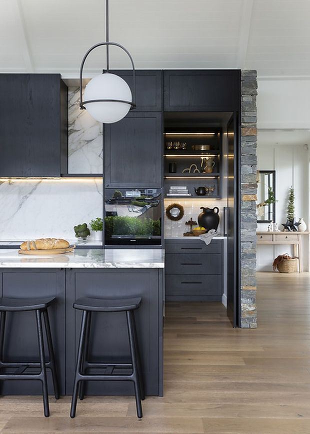 A refined contemporary kitchen with dark charcoal cabinetry, a white marble backsplash and benchtop, integrated display shelving, and a central island with two black stools. A spherical pendant light hangs above the island, while a rough stacked-stone column frames an adjoining bright living or dining area with timber flooring and built-in shelving.
