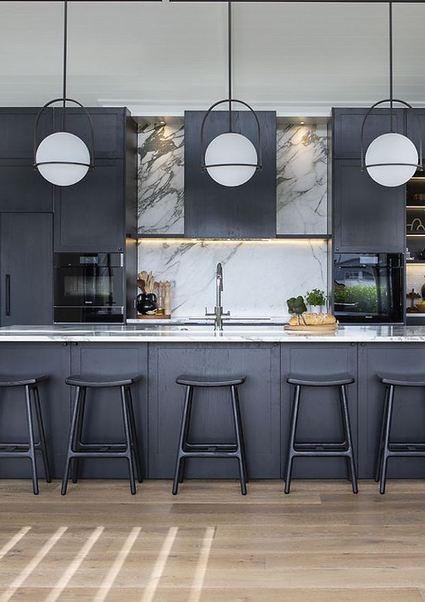 A sophisticated contemporary kitchen viewed straight on, featuring dark charcoal cabinetry, a large island with five stools, a white veined marble backsplash and benchtop, integrated appliances, and three oversized globe pendant lights. Warm timber flooring and strong natural light create contrast with the dark joinery.