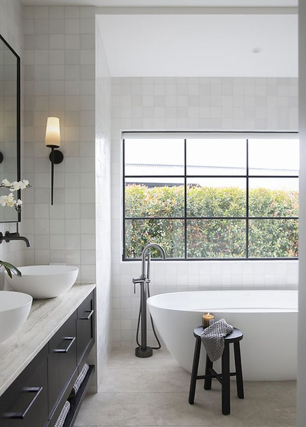 Bright contemporary bathroom with a freestanding white bathtub positioned beneath a large black-framed grid window, a double vanity with white vessel basins and dark cabinetry, pale square wall tiles, concrete-look flooring, and a minimalist black wall sconce and tapware.