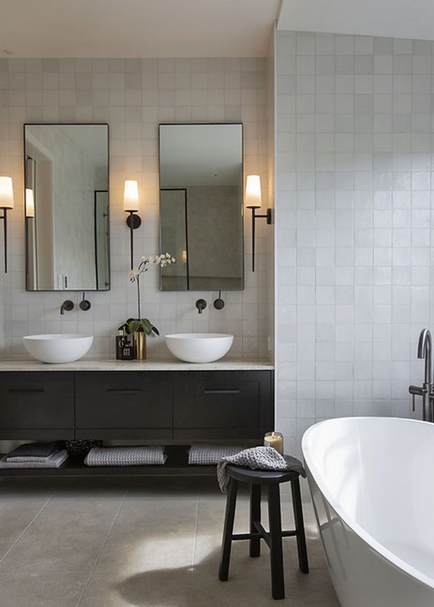 A refined contemporary bathroom with twin vessel sinks set into a dark floating vanity, two rectangular mirrors, black wall sconces, pale square tiled walls, and a freestanding white bathtub. Neutral stone-look floor tiles, black tapware, and a small stool with folded towels create a calm, monochromatic scheme.