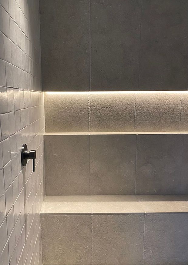 Close-up of a contemporary tiled shower or wet-room wall in muted grey tones. Large-format textured wall tiles form a recessed horizontal shelf or bench with concealed warm LED strip lighting, while smaller rectangular tiles line the side wall. A minimalist black shower control is visible.