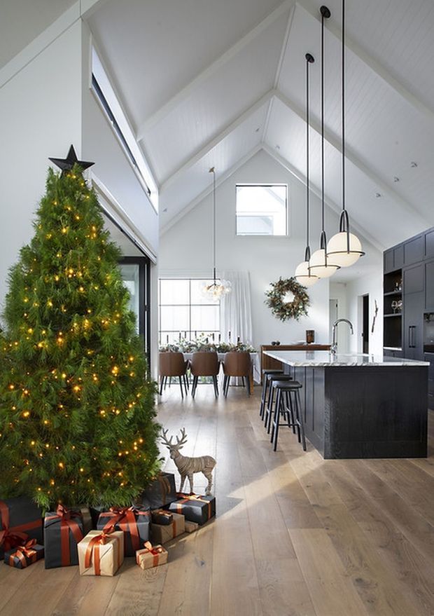 Bright open-plan residential interior with a dramatic vaulted white ceiling, exposed gable form, clerestory-style glazing and pale timber flooring. A dark kitchen island with waterfall benchtop and pendant lights occupies the right side, while a dining area sits beyond. The room is decorated for Christmas with a large lit tree, wreath, gifts and seasonal ornaments.