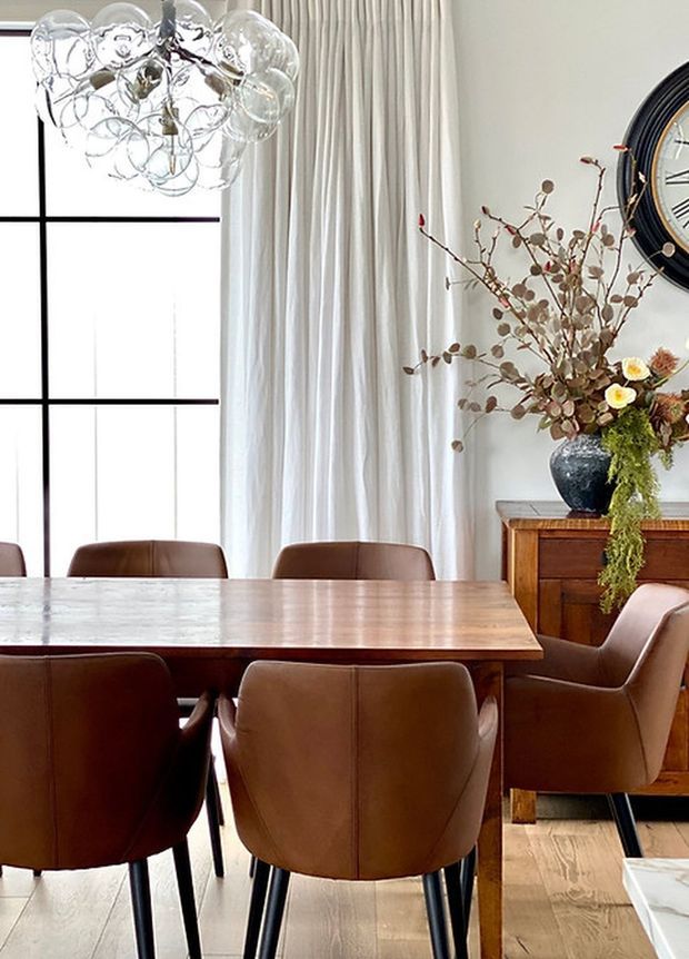 A refined dining room with a large rectangular timber dining table surrounded by brown leather upholstered chairs. The room features pale walls, full-height pleated curtains, black-framed grid glazing, light timber flooring, a sculptural clear-glass pendant light, and a wooden sideboard with decorative floral branches and a large wall clock.