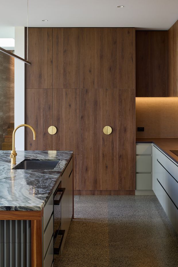 A refined contemporary kitchen with a dark natural timber wall of full-height cabinetry, pale grey handleless drawers, and a dramatic veined stone island benchtop. A brass gooseneck tap and undermount sink sit in the island, while a warm backlit small-format tiled splashback adds depth to the rear preparation area. The floor is finished in speckled terrazzo, and minimal recessed ceiling lights contribute to the clean, sophisticated composition.