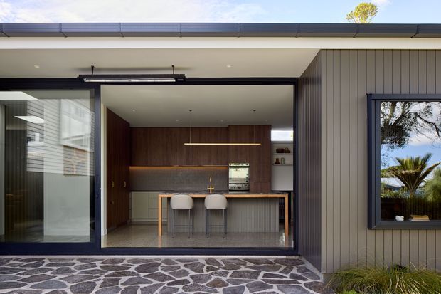 Exterior view of a contemporary single-storey home with an open, glazed kitchen entertaining area. The kitchen features dark timber cabinetry, a long island with a grey vertically ribbed front, two stools, integrated warm linear lighting, and expansive black-framed sliding glass doors. The facade uses vertical grey cladding, while the foreground patio is paved in irregular dark stone with light mortar joints. Palm planting and reflected landscaping are visible outside.