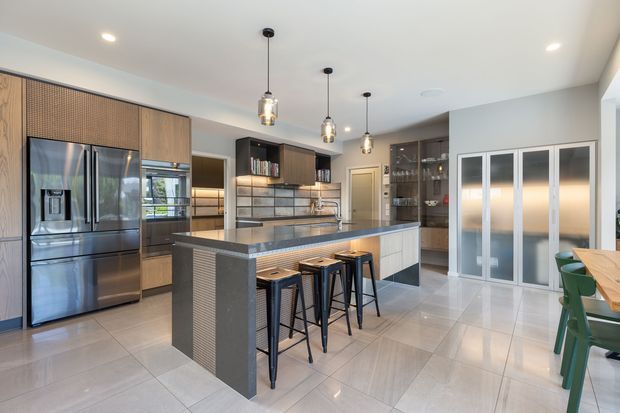 Contemporary open-plan kitchen with a large central island, integrated stainless-steel refrigerator, dark timber cabinetry, grey stone benchtops, tiled flooring, pendant lighting, and a bank of frosted-glass pantry or storage doors. A dining area is partially visible at the right.