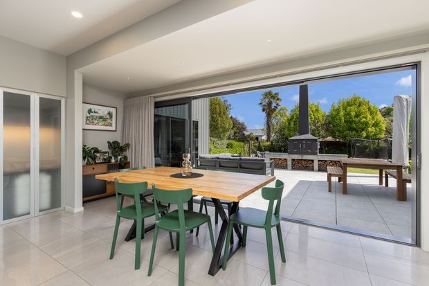 Bright contemporary open-plan dining and entertaining area with a timber dining table and green chairs, opening through expansive sliding glass doors to a tiled outdoor terrace. The room features large-format light floor tiles, white walls and ceiling, built-in cabinetry, indoor plants, and an outdoor barbecue and seating area visible beyond.
