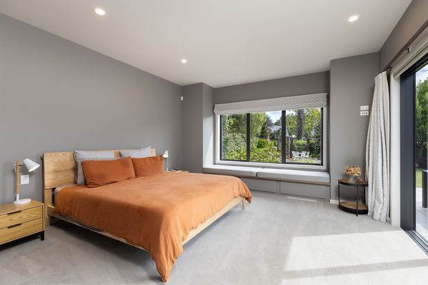 A contemporary bedroom with a low-profile timber bed, warm rust-orange bedding, grey walls, and soft light carpet. A large window seat and expansive glazed doors bring in abundant natural light and views of greenery, while a simple bedside table, lamp, and small round side table complete the room.