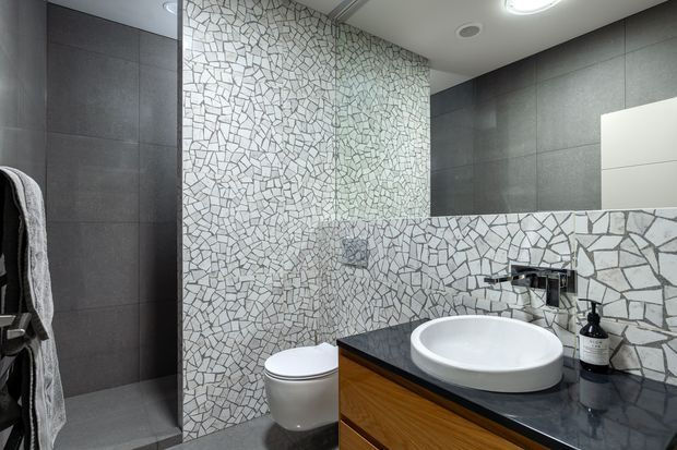 Contemporary bathroom with a wall-hung toilet, white vessel basin, dark stone-look vanity benchtop, timber vanity cabinetry, large dark grey wall tiles, and a distinctive irregular white mosaic tile feature wall and splashback. A frameless glass shower enclosure and mirror are visible.