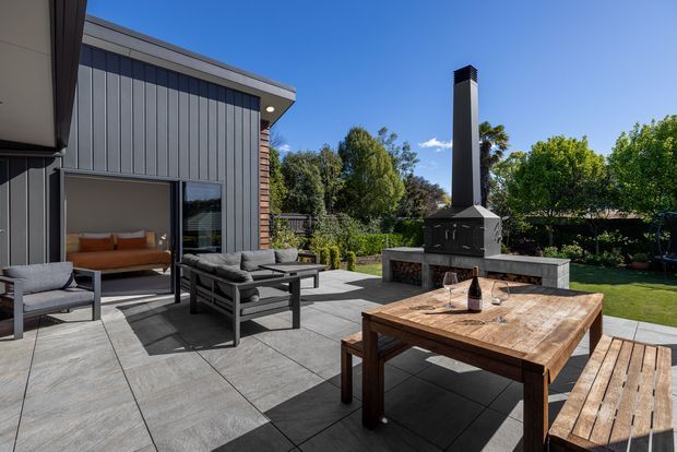 A contemporary outdoor entertaining area beside a modern home, with large grey tiled paving, a timber dining table and bench, outdoor lounge chairs, and a substantial built-in masonry fireplace with tall chimney. Dark vertical cladding, timber accents, garden planting, and a lawn surround the terrace; an interior living space is visible through the glazed door.