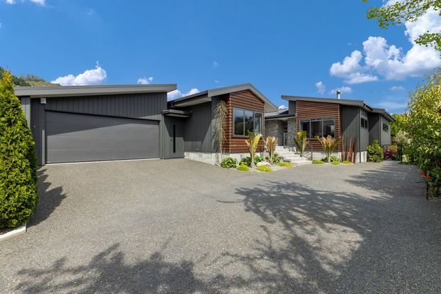 Exterior view of a contemporary single-storey residential home with a dark grey attached garage and vertical cladding, complemented by timber-look facade panels around the entry and front windows. A broad gravel driveway and simple landscaped planting lead to the entrance under a bright blue sky.