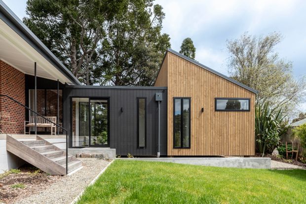Exterior view of a contemporary single-storey extension connecting to an existing red-brick house. The addition has a simple pitched/gable roof, vertical timber cladding on the main volume, dark vertical cladding on the link section, black-framed glazing, and a raised concrete base. A covered entry deck and steps are visible at left, with lawn and established trees around the building.