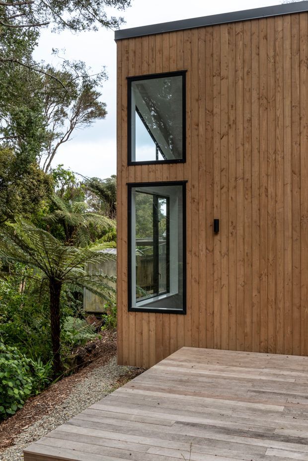 Exterior view of a compact modern building in a lush wooded setting. The facade is clad in vertical warm timber boards with slim black metal window frames and a dark roof edge. Two tall windows reflect surrounding trees, while a broad timber deck occupies the foreground and gravel landscaping borders the structure.