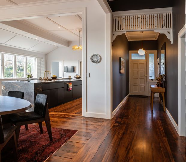 Interior view of a refined home showing a dark timber-floored hallway and entry leading into an open kitchen and dining area. The kitchen has dark cabinetry, a pale stone island benchtop, and large windows with plantation shutters. The hallway features charcoal walls, white decorative architraves, a pendant light, a timber console, and an ornate white balustrade over the opening, creating a strong contrast between traditional detailing and contemporary furnishings.