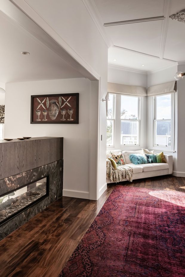 A refined living room interior with a white-painted, heavily moulded coffered ceiling and bay window seating area. The room features dark timber flooring, a contemporary linear fireplace clad in dark veined stone, a neutral sofa with colourful cushions and a throw, and a large richly patterned burgundy rug. Decorative wall art and classic trim details create a polished traditional character.