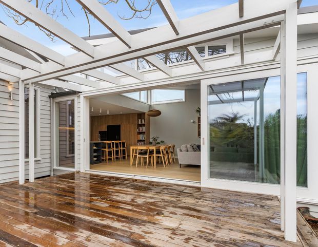 A covered outdoor timber deck and courtyard-like terrace viewed toward an open-plan living and dining area. White-painted pergola beams and posts frame large sliding glass doors, with white weatherboard walls and a partially enclosed roof structure. Inside, a timber dining table and chairs, sofa, television, shelving, and warm wood finishes are visible. The wet deck reflects the overcast daylight, while climbing vines and trees appear above and through the glazing.