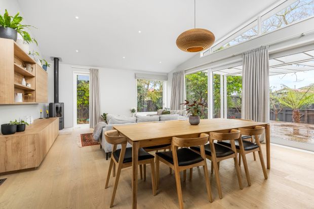 Bright open-plan dining and living area with a long solid-wood dining table and matching timber chairs, integrated timber cabinetry and shelving, pale timber flooring, white walls and ceiling, expansive floor-to-ceiling glazing with sheer curtains, and a sculptural woven timber pendant light. A small seating area and fireplace are visible behind the dining zone, with greenery outside.