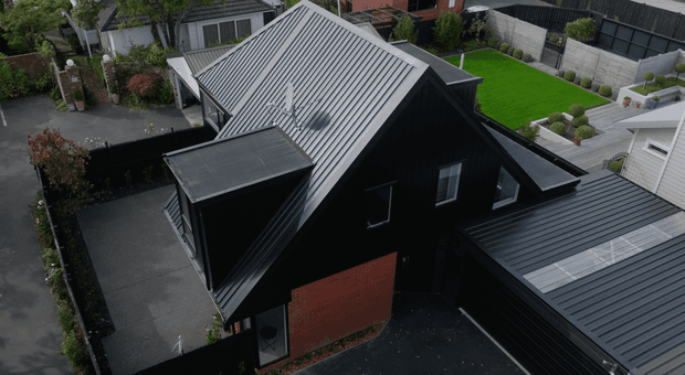 Elevated aerial view of a contemporary dark-clad residential house with a steep gabled metal roof, black vertical cladding, and a contrasting red brick wall. Multiple roof forms and extensions surround paved driveways and landscaped suburban properties.
