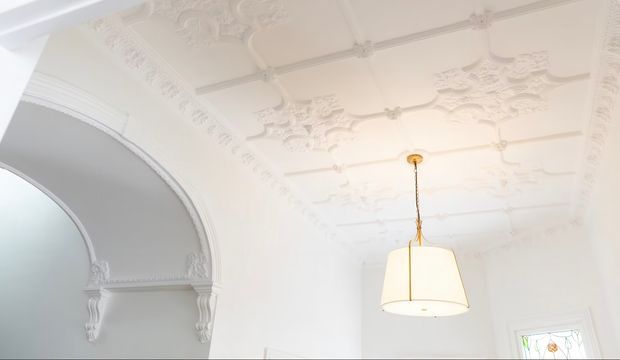 Bright white interior featuring an ornate decorative plaster ceiling with raised floral panels, moulded cornices and ceiling roses. A large arched opening with intricate moulded trim and decorative corbels is visible on the left, while a simple pendant light hangs centrally. A small leadlight-style window appears at the lower right.
