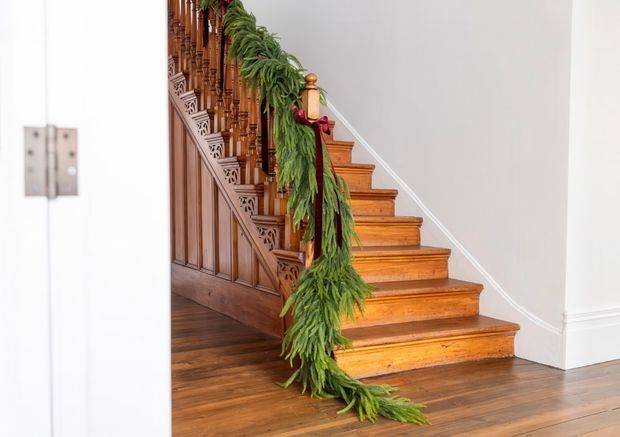 A bright interior view of a traditional timber staircase with richly detailed carved balusters, paneled stringer, turned newel post, and polished wooden treads. The staircase rises against white walls and white trim, with a green garland draped along the banister. Dark timber flooring continues through the hall.