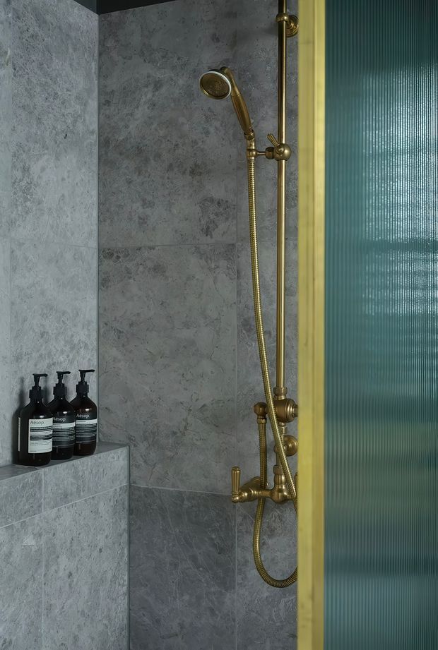 Close-up of a refined shower enclosure with grey veined stone-look wall tiles, a built-in tiled ledge holding black toiletry bottles, and brushed brass/gold shower fixtures including a handheld shower, rail, hose, and mixer. A vertically ribbed translucent glass door or screen with a brass-toned frame is visible on the right. The lighting is moody and the palette is predominantly grey with metallic gold accents.