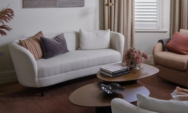 A refined living room with a white upholstered sofa, layered neutral and muted mauve cushions, a textured beige armchair, and overlapping rounded timber coffee tables. The room features warm brown flooring, full-length beige curtains, white plantation shutters, a simple wall-mounted textured artwork, and soft floral styling, creating a calm, warm, contemporary-classic atmosphere.