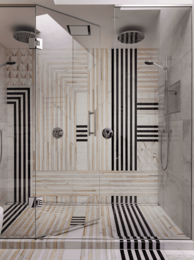 A contemporary bathroom dominated by a large glass-enclosed walk-in shower. The shower walls and floor feature pale stone or marble-look surfaces with linear veining, geometric herringbone sections, and bold black-and-white striped tile accents. Multiple overhead rain showerheads, wall-mounted controls, and handheld shower fittings are visible, with mirrored glass amplifying the space. The overall look is highly graphic, monochromatic, and refined.