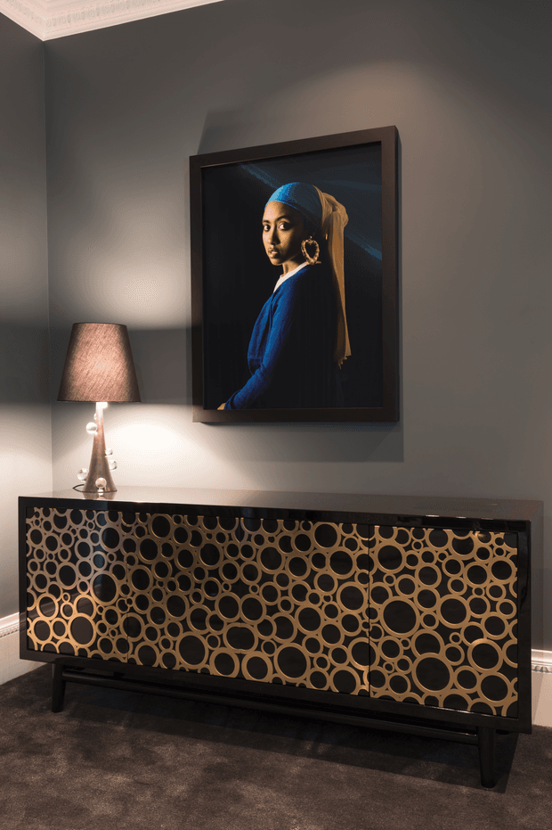A moody, refined interior vignette featuring a dark grey wall with ornate white ceiling cornice, a framed portrait, and a long black sideboard with striking circular patterned cabinet fronts in warm gold and brown tones. A sculptural table lamp provides warm accent lighting, while dark carpet and black furniture legs reinforce the dramatic palette.