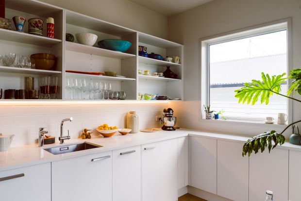 Bright, compact contemporary kitchen with white flat-panel cabinetry, white tiled backsplash, pale benchtops, open display shelving, integrated under-shelf lighting, stainless-steel sink and tap, and a large window. The shelving displays glassware and colourful crockery, while indoor plants add a natural accent.