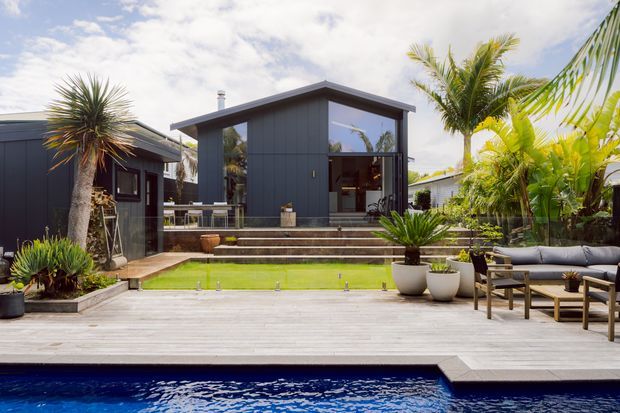 Contemporary dark-clad single-storey home viewed from a landscaped outdoor entertaining area. The house features a simple low-pitched roof, large glazed openings, broad timber decking, stepped paving, lush tropical planting, and a swimming pool in the foreground. Outdoor seating occupies the right side of the deck.