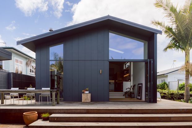 A compact contemporary dark-clad pavilion or backyard extension with a low-pitched roof, large glazed openings, and wide timber decking with stepped access. The open glazed doorway reveals a warm interior, while palm trees and neighbouring lightweight homes suggest a sunny suburban or coastal setting.