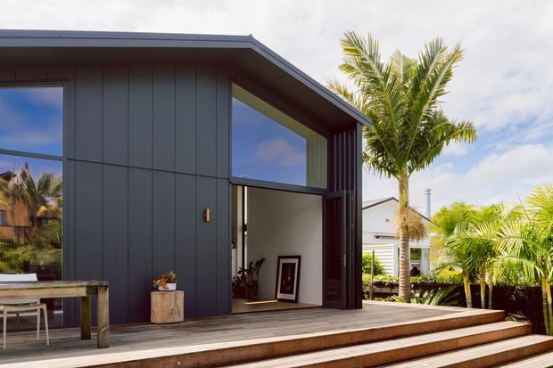 Exterior view of a compact contemporary pavilion or extension with dark vertical cladding, a low mono-pitched roof, large glazed openings, and broad timber steps leading to an open entry. Tropical palms and a neighbouring light-coloured building are visible around the structure.