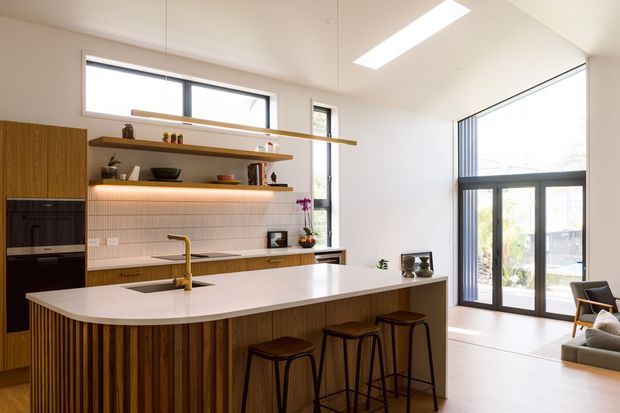 Bright contemporary kitchen with a large island and integrated seating, timber veneer cabinetry, open timber shelving, a pale benchtop, and a tiled splashback. Full-height glazing and a skylight bring abundant natural light, with a small lounge area visible beyond the kitchen.