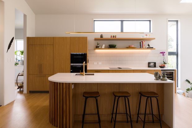 Bright, contemporary kitchen with a large island featuring a white stone benchtop, vertically fluted timber cabinetry, integrated appliances, open timber shelves, and a pale pink tiled splashback. Three black stools line the island, while warm timber flooring and understated indoor plants add warmth. Tall windows provide abundant natural light.
