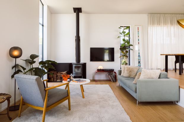 Bright, warm contemporary living room with a pale grey sectional sofa, mid-century-style lounge chair, timber flooring, white walls, a large light rug, indoor plants, wall-mounted television, and a black wood-burning stove with exposed flue. Full-height glazing and sheer curtains bring in abundant natural light, while a dining area is visible beyond the living space.