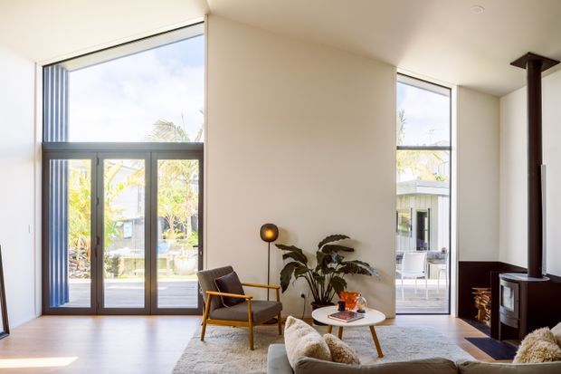 Bright contemporary living room with expansive floor-to-ceiling glazing and glazed doors, a pale timber floor, cream walls, and a central column. Mid-century-inspired lounge chairs, a low round coffee table, a large neutral rug, indoor plants, and a freestanding dark wood-burning stove create a warm, relaxed interior. The outdoor garden and neighbouring buildings are visible through the glazing.