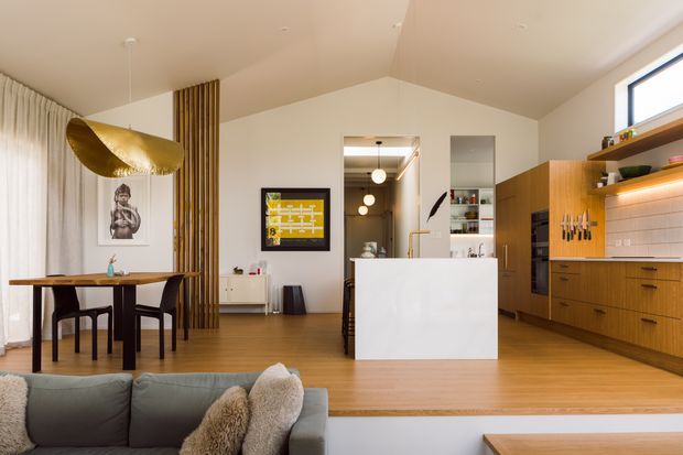 Open-plan interior showing a warm contemporary kitchen and dining area, viewed from the living room. The kitchen features a white island, extensive timber cabinetry, open shelving with integrated lighting, a tiled backsplash, and dark-framed glazing. A timber dining table sits beside a sculptural gold pendant light, with vertical timber screening separating the dining zone from the kitchen. The room has light wood flooring, white walls and ceiling, and a raised split-level transition to the living area.