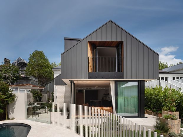 A contemporary multi-storey residential house viewed from the street, featuring a steep front-facing gable roof, dark vertically panelled cladding, a recessed upper balcony, and a partially open ground-floor entertaining area. Large glazing, light brickwork, timber soffit detailing, and a white vertical-slat boundary fence define the facade. A small swimming pool and landscaped planting are visible in the foreground.