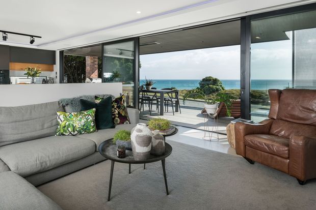Bright contemporary open-plan living room with a large grey sectional sofa, brown leather armchair, round coffee tables, and floor-to-ceiling sliding glass doors opening to a balcony or terrace with ocean views. The space includes a partially visible modern kitchen, polished concrete or light flooring, dark-framed glazing, and a neutral palette accented by indoor plants and patterned cushions.