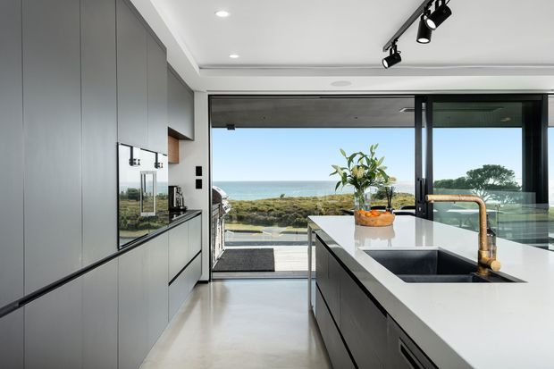 A sleek contemporary kitchen with floor-to-ceiling glazing and expansive ocean views. The room features streamlined matte grey cabinetry, integrated appliances, a large white island with a dark inset sink, and a warm brass-toned tap. Track lighting and recessed ceiling lights provide a clean, modern finish.