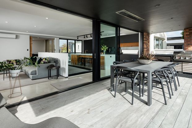 Contemporary indoor-outdoor entertaining area with a covered patio deck furnished with a large dining table and chairs. Full-height black-framed sliding glass doors open to a bright open-plan living and kitchen area, with a visible kitchen island, timber accents, exposed brick, and neutral furnishings. The deck has pale timber-look flooring and a built-in outdoor kitchen or barbecue zone at the right.