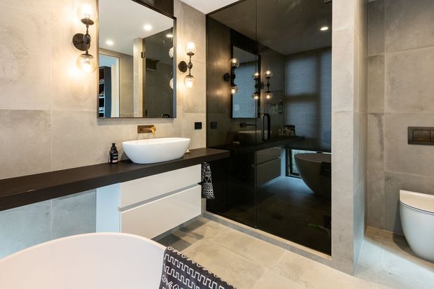 Contemporary bathroom with a floating white vanity, dark benchtop, vessel basin, large mirrored wall, warm globe sconces, and a glass-enclosed dark tiled shower area. A freestanding bathtub is visible in the foreground, with a toilet partially visible in a separate nook. The palette combines pale stone-look wall and floor tiles with charcoal cabinetry and dark feature surfaces.