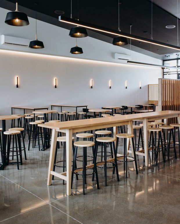 A bright contemporary commercial dining or café interior with rows of communal timber tables and black metal stools. The space features polished concrete flooring, white walls, black ceilings, linear pendant lighting, black dome pendant lights, slim wall sconces, exposed air-conditioning units, and a timber slatted privacy screen. The open, orderly layout has an industrial-modern character.