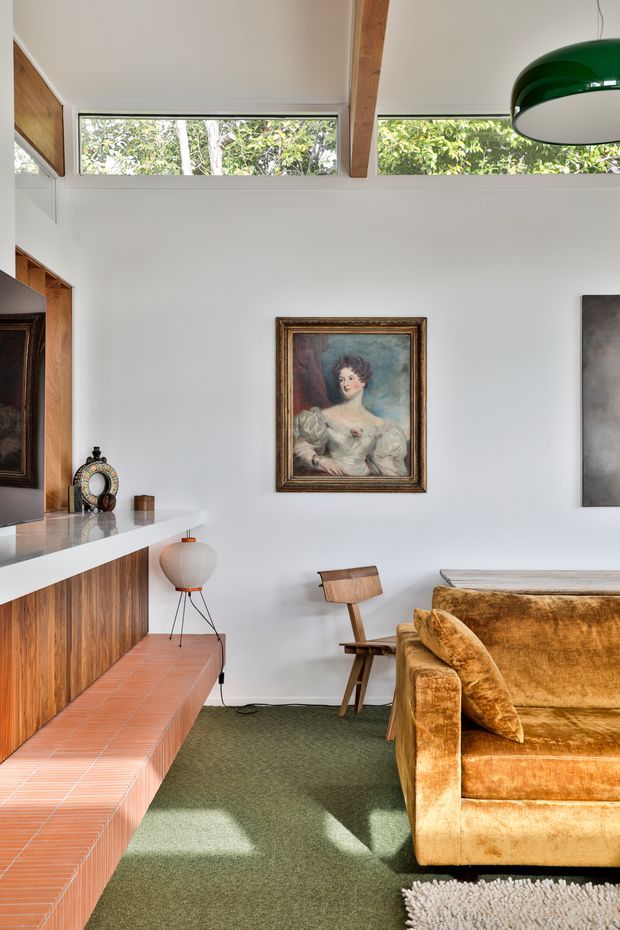Bright, characterful living room with a white-painted interior, high clerestory windows, exposed timber beam, and warm timber wall panelling. A large white built-in ledge and terracotta-tiled hearth or bench run along the left wall, with a vintage framed portrait, sculptural pendant light, mid-century wooden chair, and plush ochre velvet sofa. Green carpet and abundant daylight create a warm eclectic atmosphere.