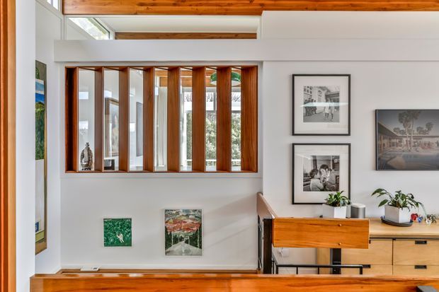 Interior view of a bright, mid-century-inspired living or circulation space with white walls, exposed timber ceiling beams, warm timber flooring and joinery, and a distinctive internal timber-framed glazed partition. Framed artwork, a timber console, cabinetry, and indoor plants furnish the room. Clerestory glazing and the elevated ceiling bring in natural light.